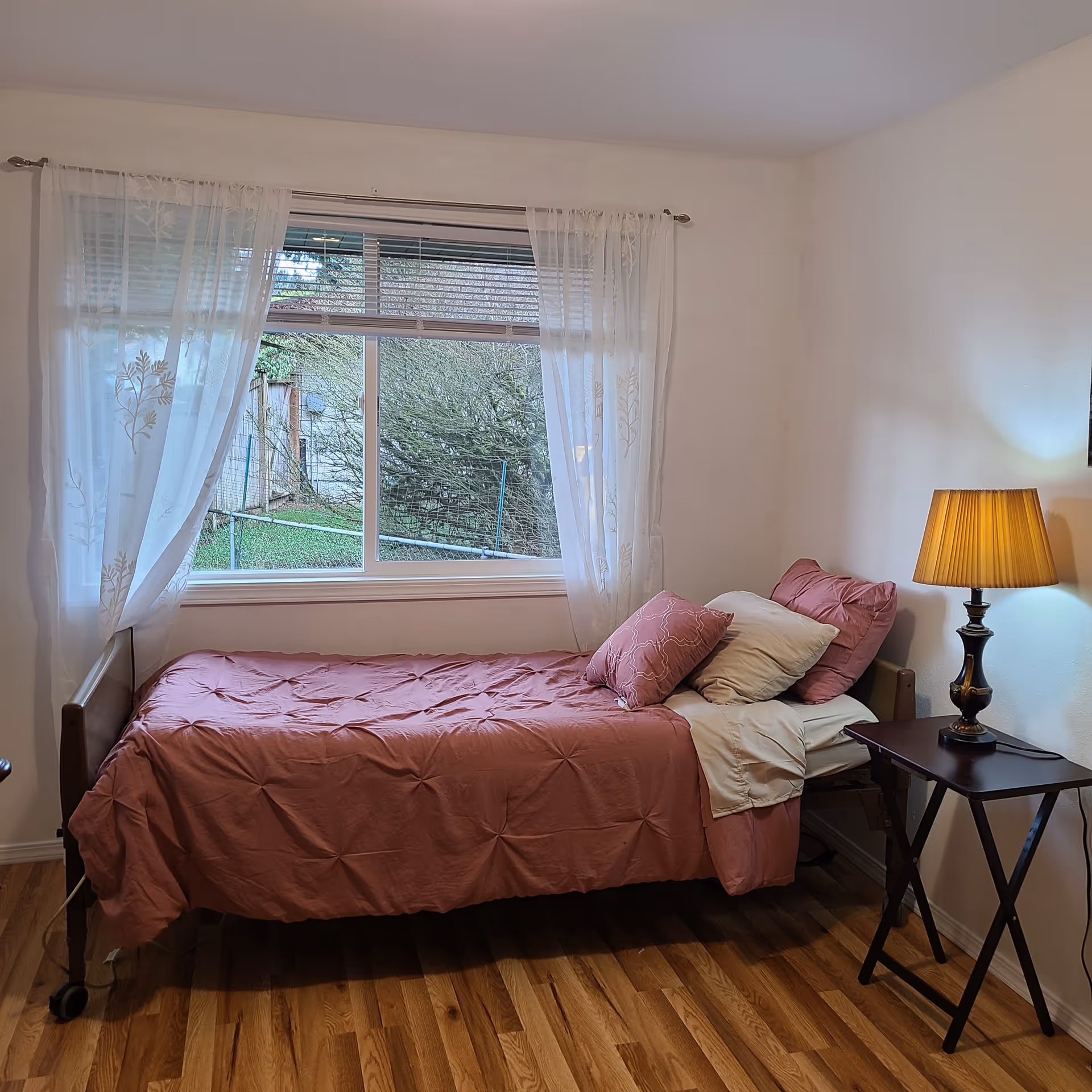 A small furnished bedroom with a pink quilt, window with sheer curtains, and a side lamp.