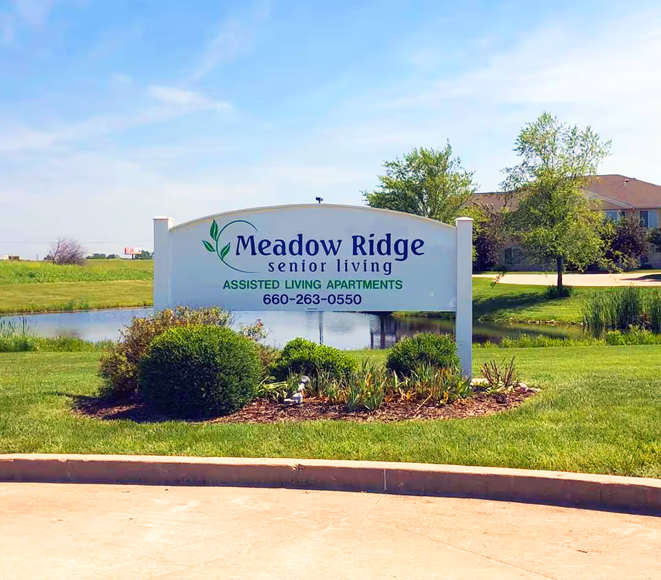 Outdoor view of a Meadow Ridge Senior Living sign with green bushes and a small pond behind it, under a clear blue sky.