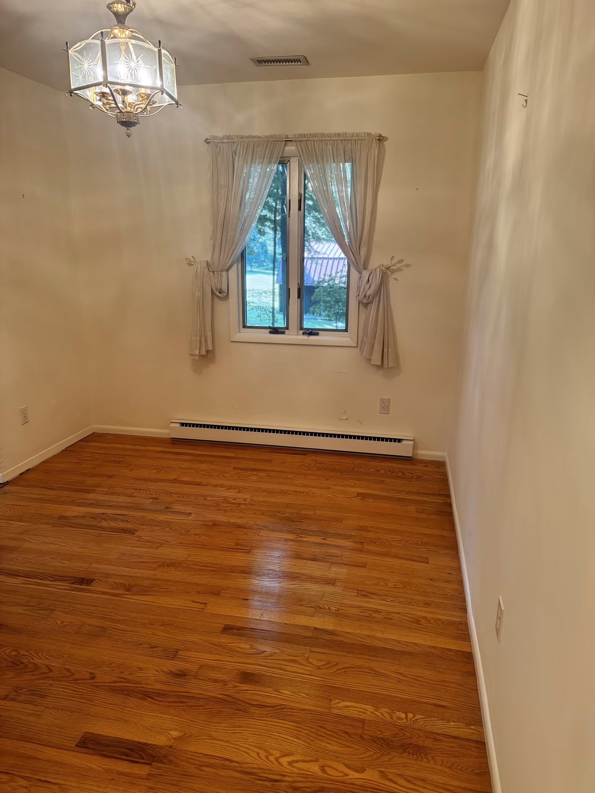 Empty small room with hardwood floor, window with curtains, and a hanging ceiling light.