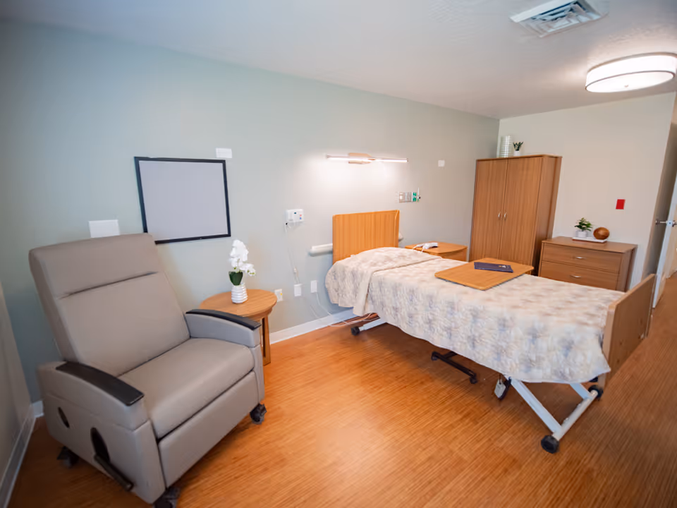 A patient bedroom in a senior care center with a bed, recliner, and wooden furniture.