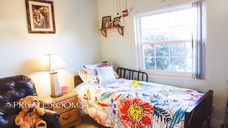 Small private bedroom with a twin bed covered in a colorful floral bedspread, a nightstand and lamp, a leather chair with a stuffed bear, and a window.