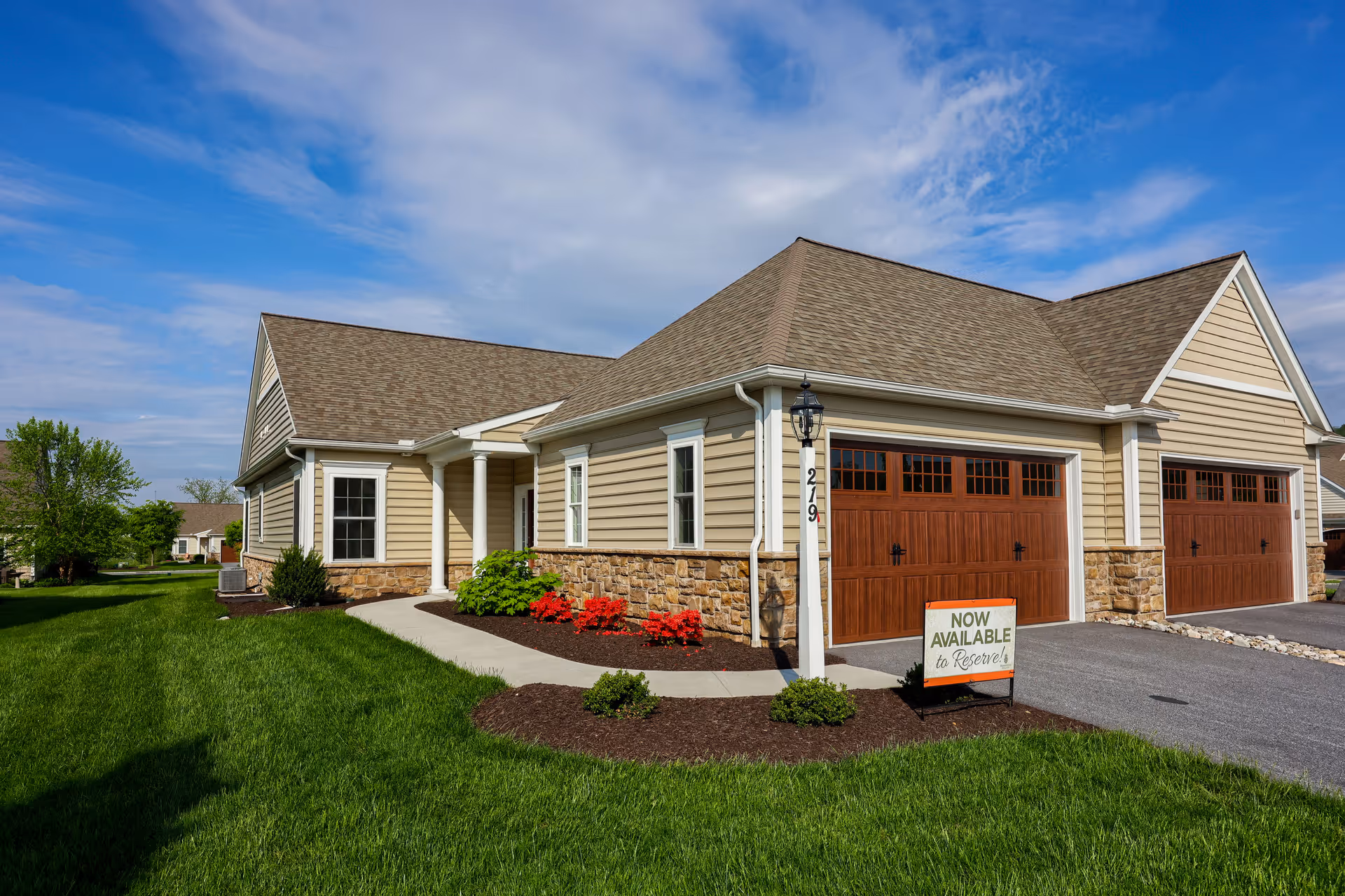 Front exterior of a single-story Homestead Village residence with two wooden garage doors, manicured lawn, and a "Now Available" sign.