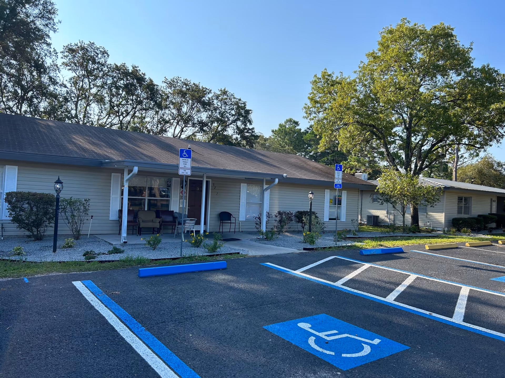 Exterior view of a single-story senior living facility building with a covered porch area featuring chairs. There are two handicap parking spaces marked in blue in front of the building. Trees and shrubs surround the building under a clear blue sky.