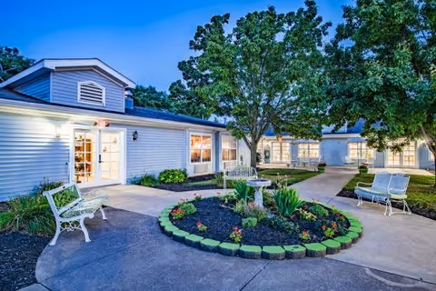 Outdoor courtyard area at a senior living facility with white benches, a circular flower bed with plants and flowers, paved walkways, and single-story buildings with white siding and large windows under a clear evening sky.
