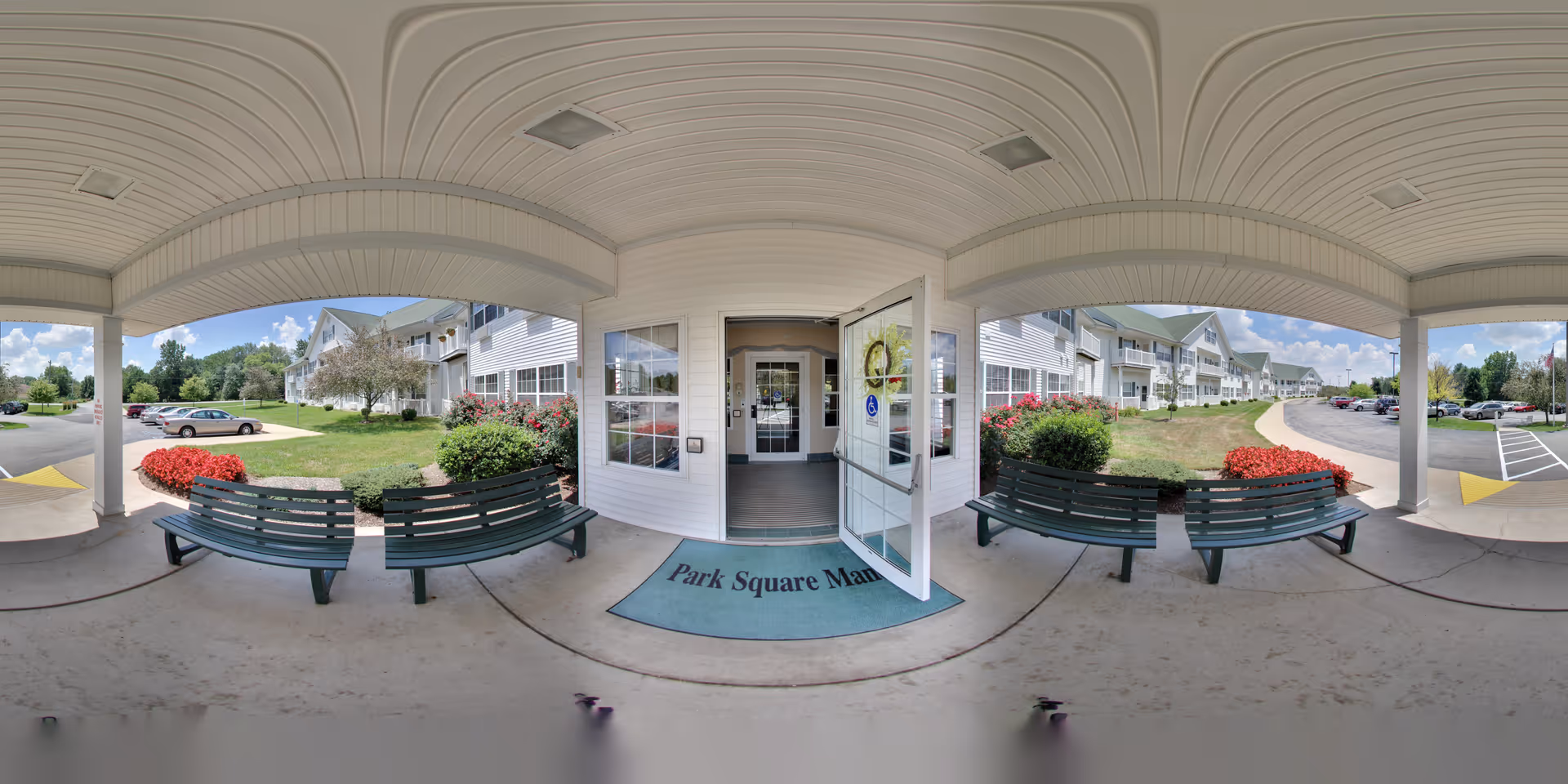Covered entrance area of Park Square Manor with two green benches facing each other, a blue welcome mat with the facility name, and an open glass door leading inside. The exterior of the building is white with multiple windows and a well-maintained lawn with red flowers and bushes. A parking lot with several cars is visible on both sides under a partly cloudy sky.