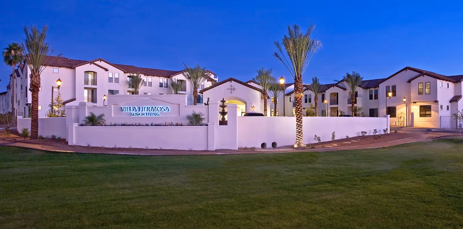 Dusk exterior view of the Villa Hermosa Senior Living complex with an illuminated entrance wall, palm trees, and landscaped lawn.
