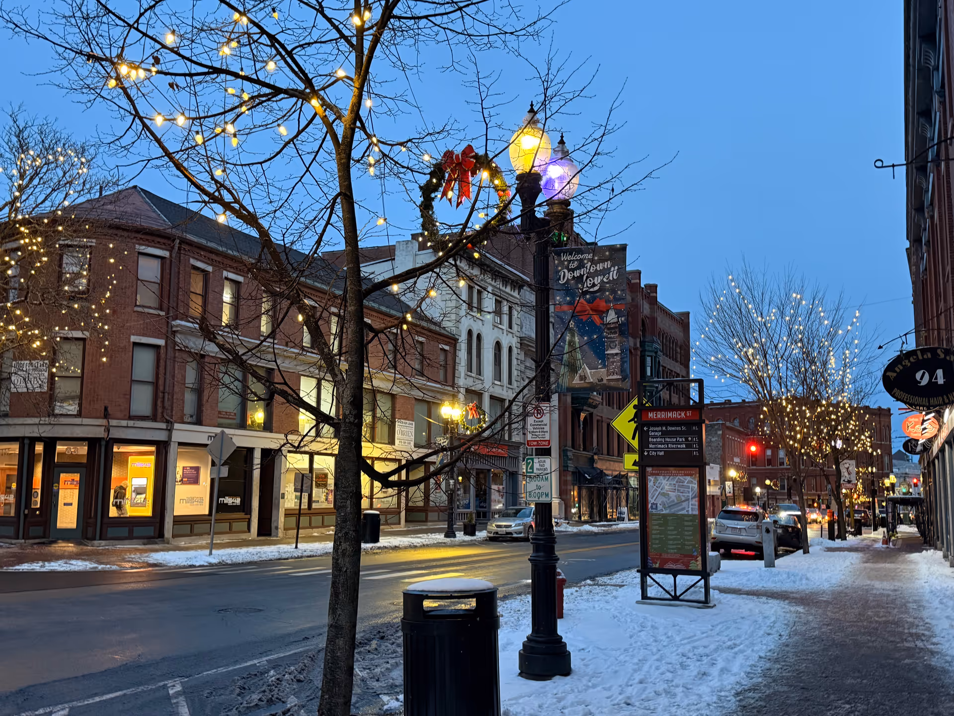 Snowy downtown street with festive lights and storefronts along a brick-lined avenue at dusk.
