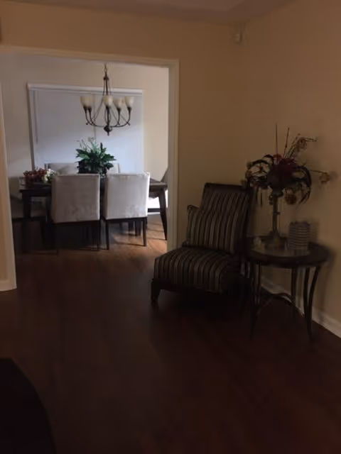 Interior view showing a small sitting area with a striped armchair and a round side table with a floral arrangement. In the background, there is a dining area with a dark wooden table, six light-colored upholstered chairs, a chandelier overhead, and a large window with closed blinds.