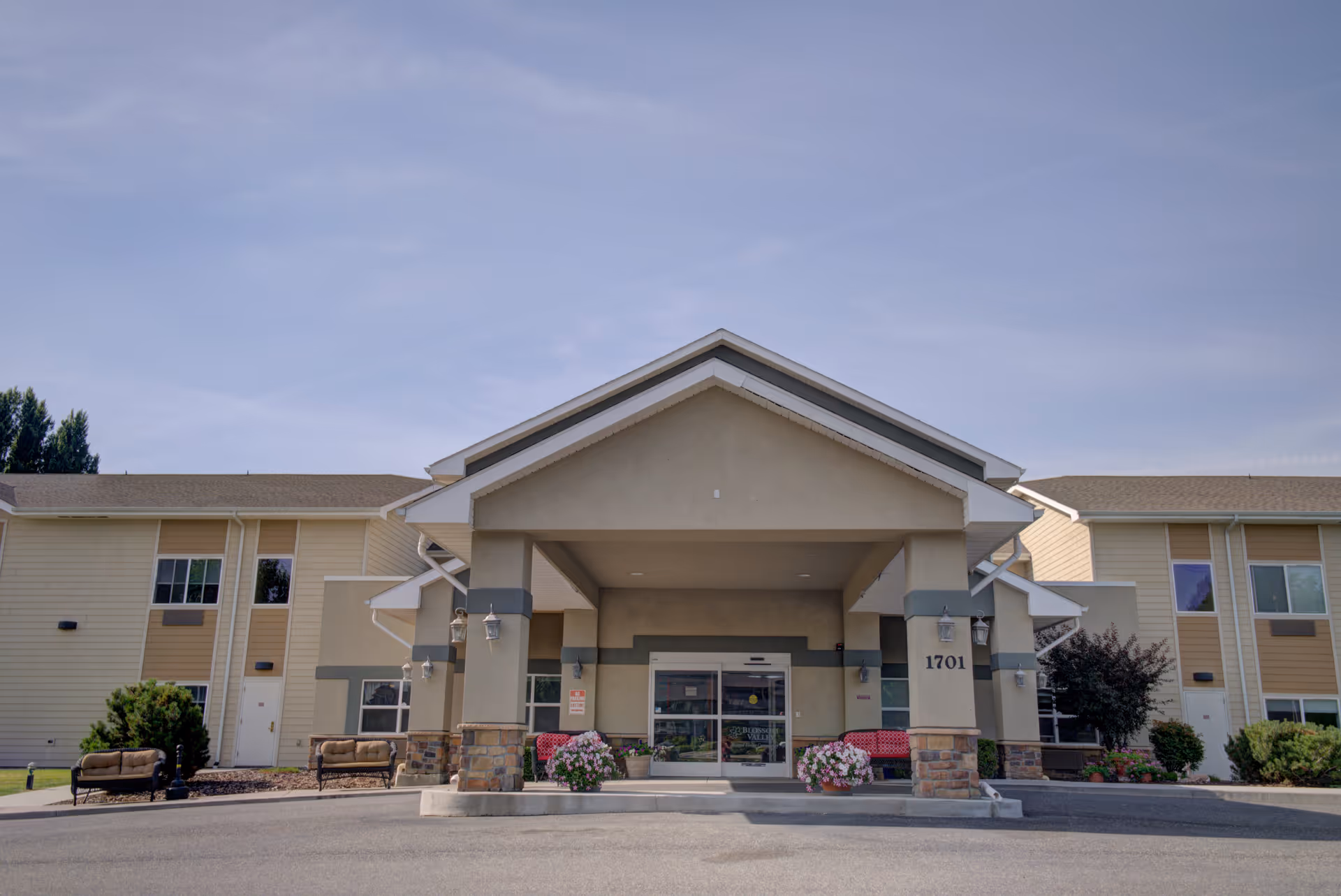 Front exterior view of a senior living facility building with a covered entrance, two benches on either side, flower pots near the entrance, and the address number 1701 displayed on a pillar.
