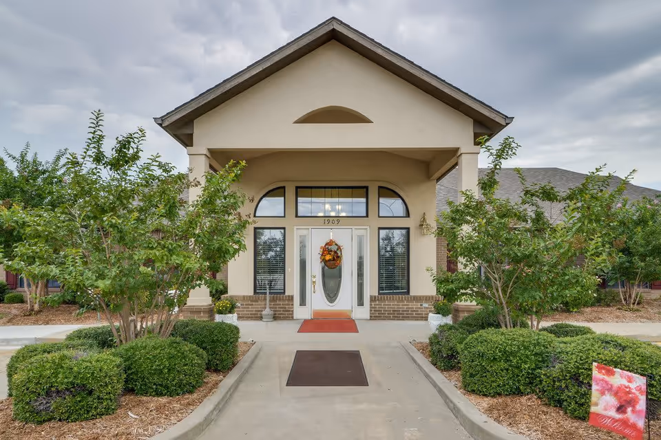 Front exterior view of a senior living facility entrance with a covered porch, a glass door decorated with a fall wreath, surrounded by windows and landscaped with bushes and small trees.