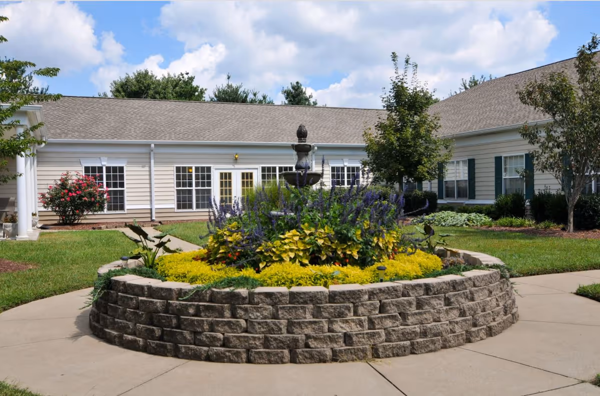 Circular stone planter with flowers and fountain sits in front of a single-story senior living building.