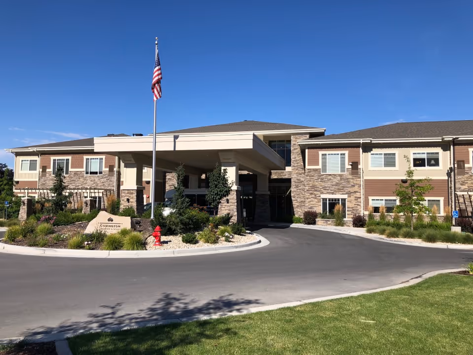 Exterior view of a senior living facility building with a covered entrance, stone and siding facade, an American flag on a flagpole, landscaped greenery, and a driveway in front.