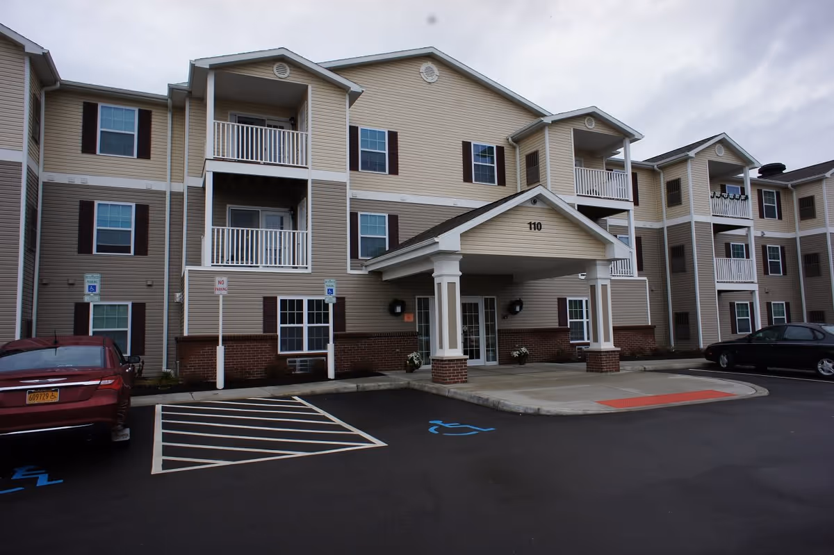 Exterior view of a three-story beige and brick residential building with balconies and a covered entrance marked with the number 110. There are two cars parked in front, including a red car in a handicapped parking space. The sky is overcast.