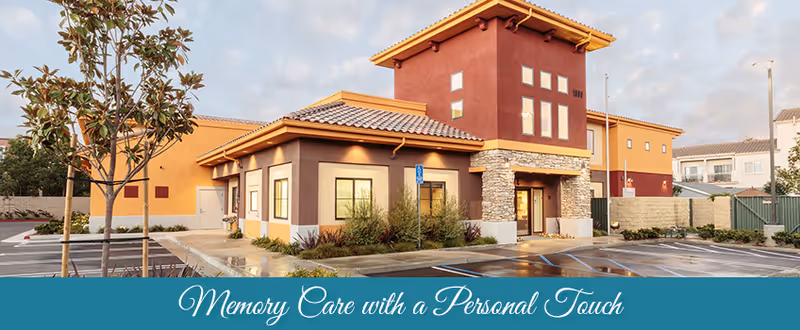 Exterior view of a modern memory care facility building with a combination of tan, brown, and red colors, stone accents, and a tiled roof. The parking lot in front has marked spaces including handicapped spots, and there are small trees and landscaping around the building. The sky is partly cloudy during daylight.
