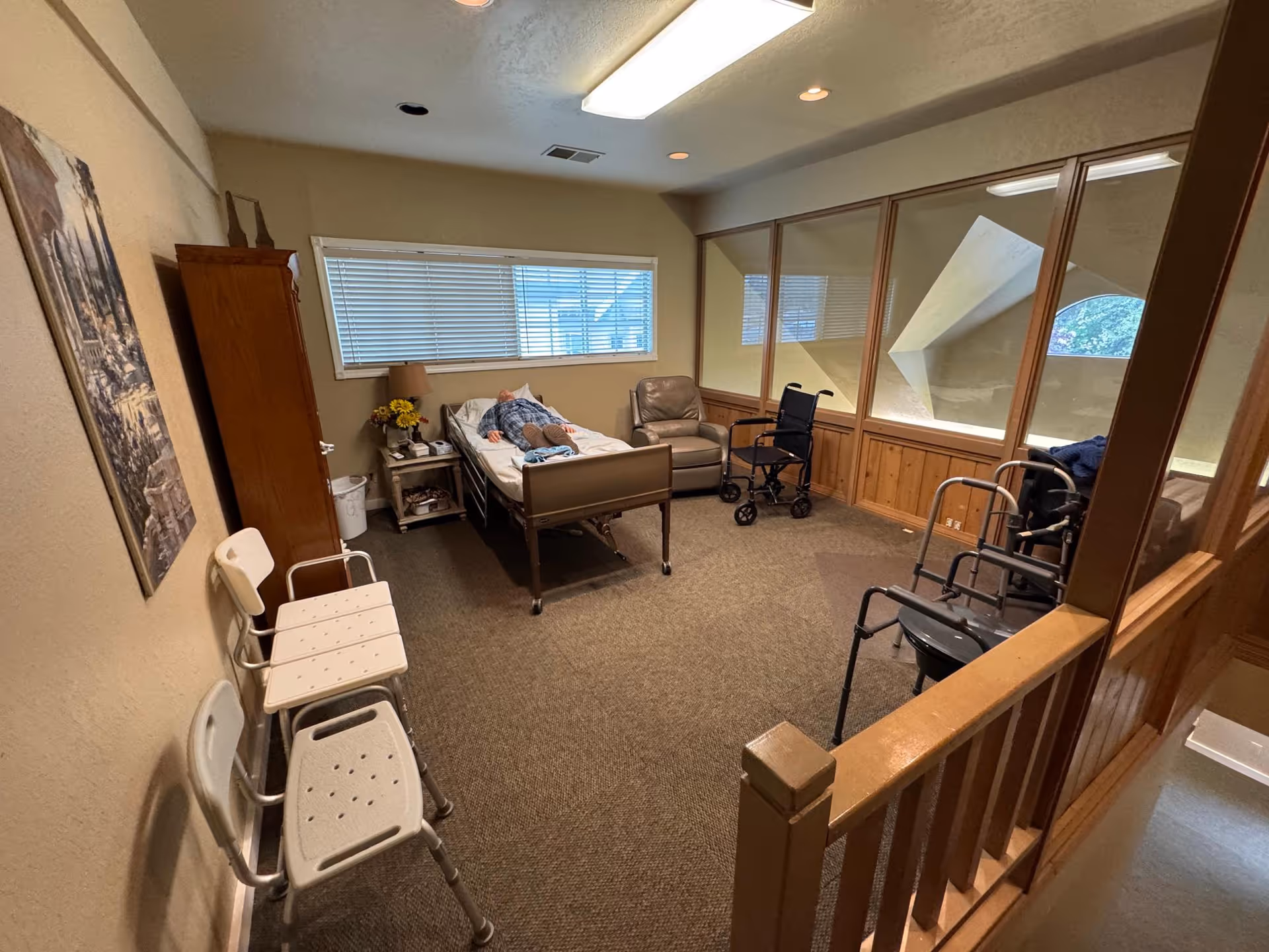 A senior care facility interior featuring a patient in bed with chairs, walker, and wood paneling.
