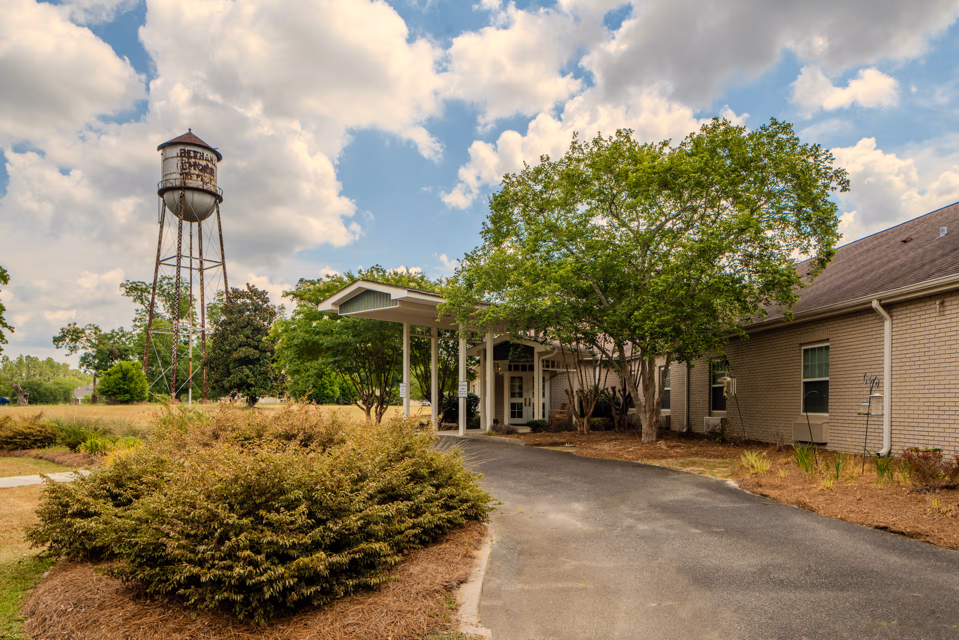 Exterior view of The Oaks Bethany Assisted Living, featuring the entrance, driveway, trees, and water tower.