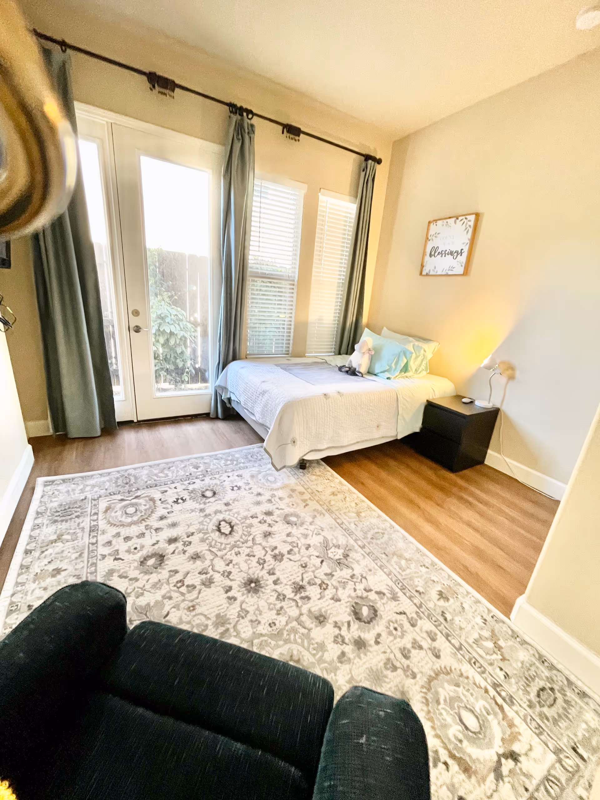 A bright bedroom with a single bed against the wall, decorated with light blue pillows and a stuffed animal. There is a black nightstand with a lamp next to the bed. The room has large windows and a glass door with blue curtains, letting in natural light. A patterned area rug covers the wooden floor, and a black armchair is partially visible in the foreground. A framed sign with the word 'blessings' hangs on the wall above the bed.