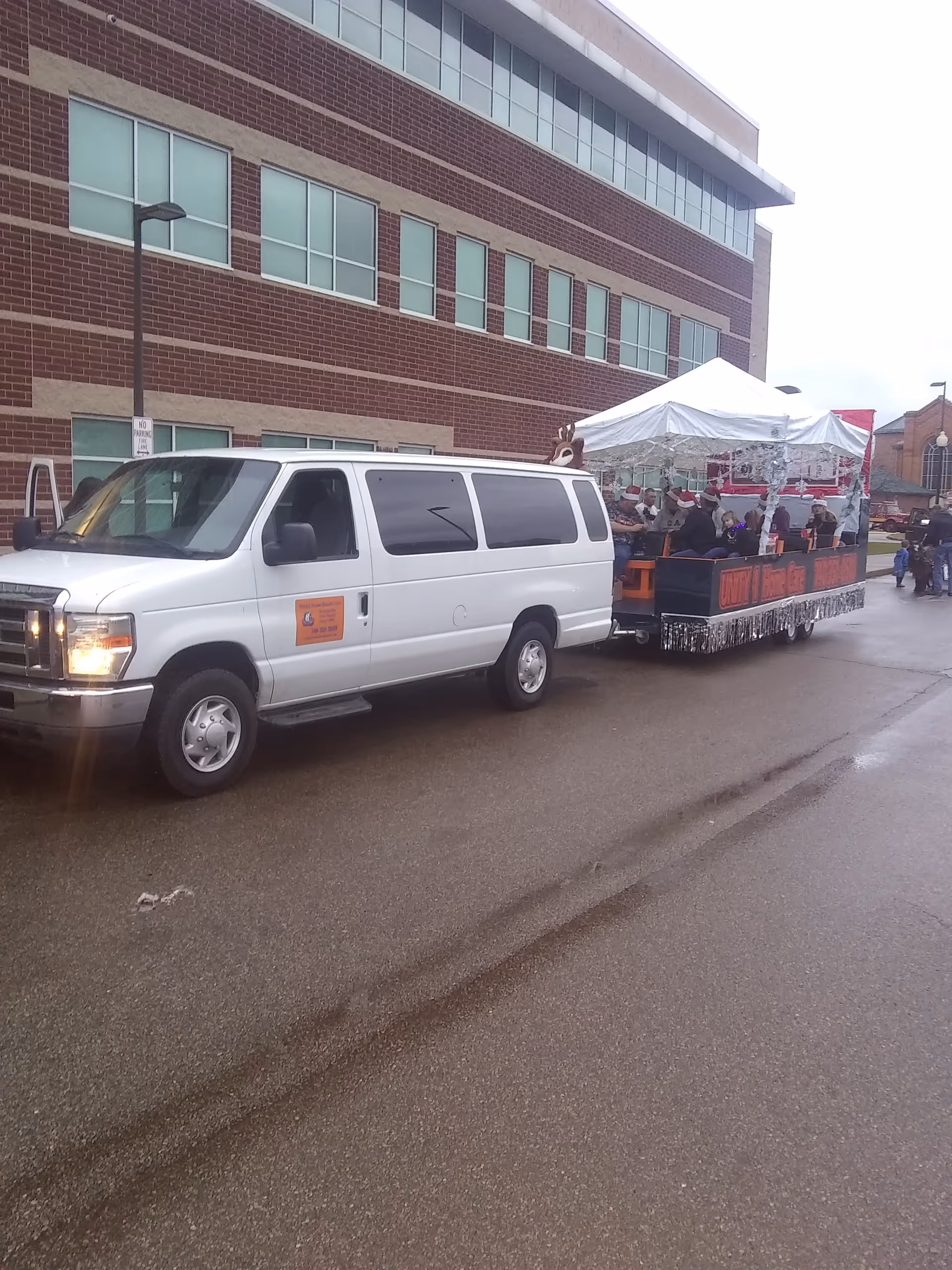 White van pulling a decorated parade float with people along a brick-building street.