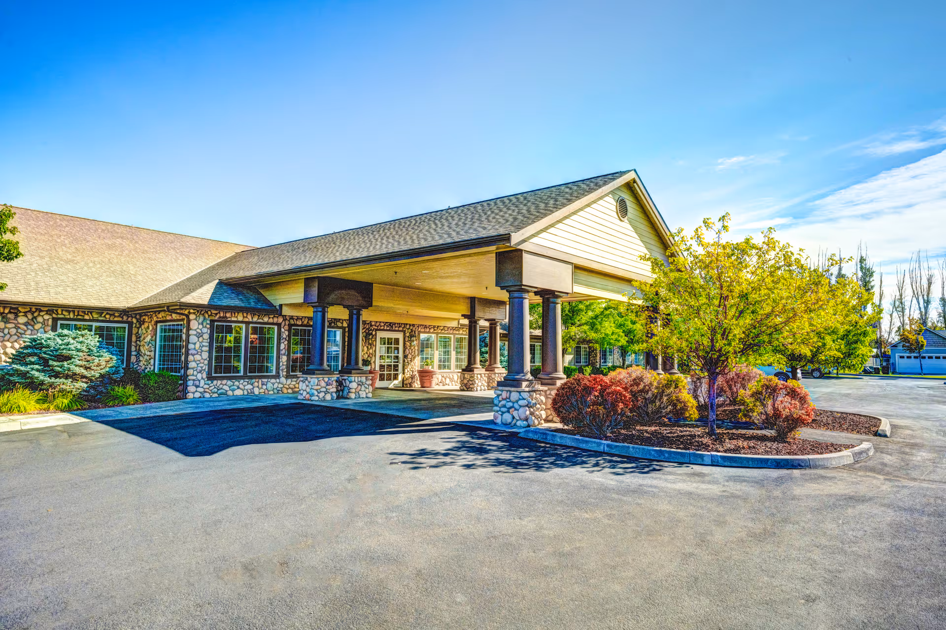Exterior view of a single-story building with a stone facade and a covered entrance supported by columns. There is a landscaped area with trees and shrubs near the entrance, and a clear blue sky overhead.