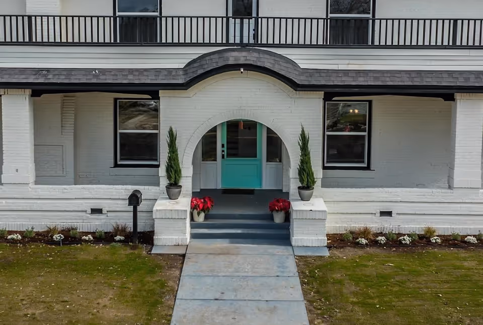 White brick building with a curved entry arch and teal front door flanked by potted evergreens.