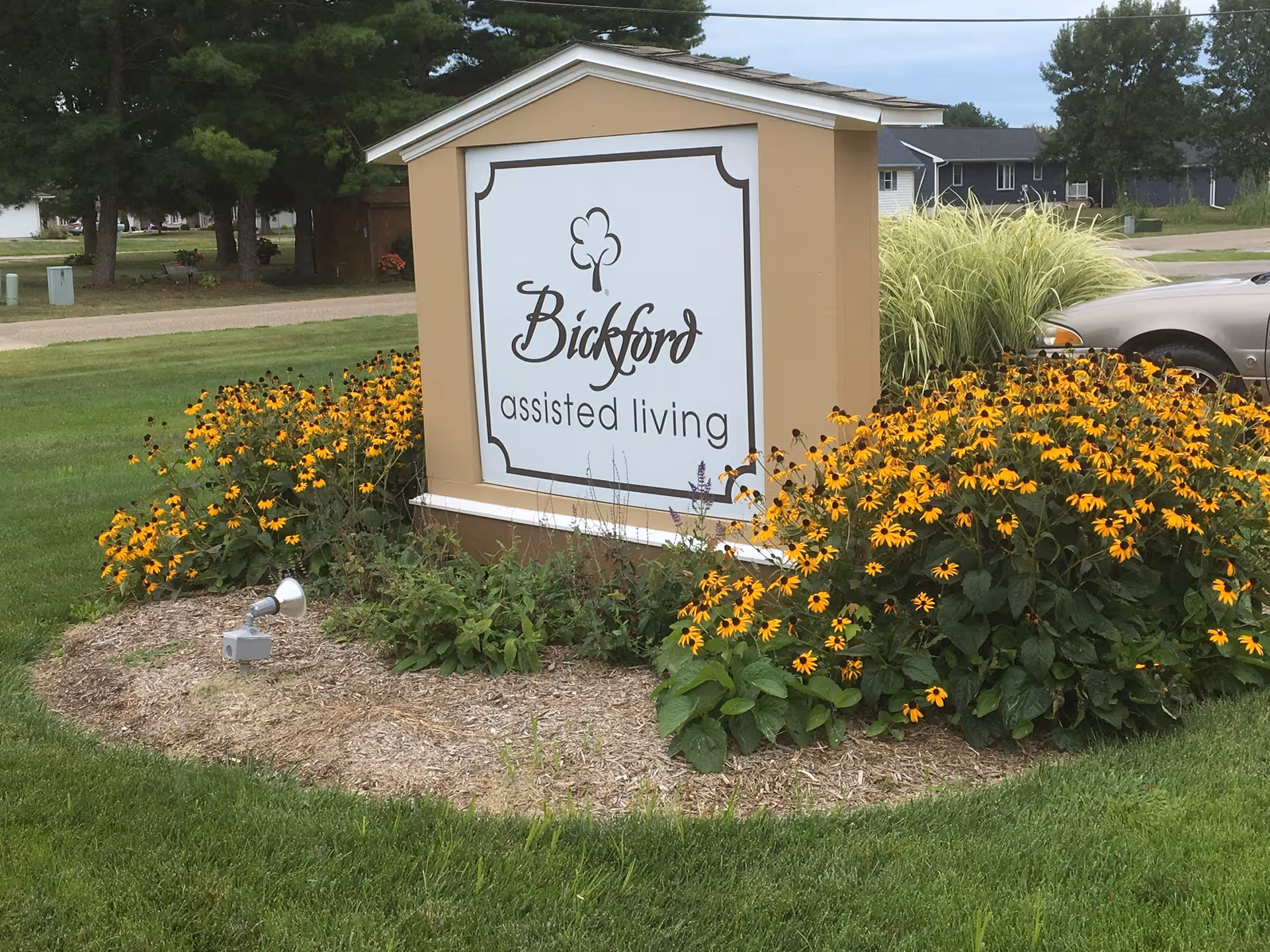 Outdoor sign reading 'Bickford assisted living' surrounded by vibrant yellow flowers.