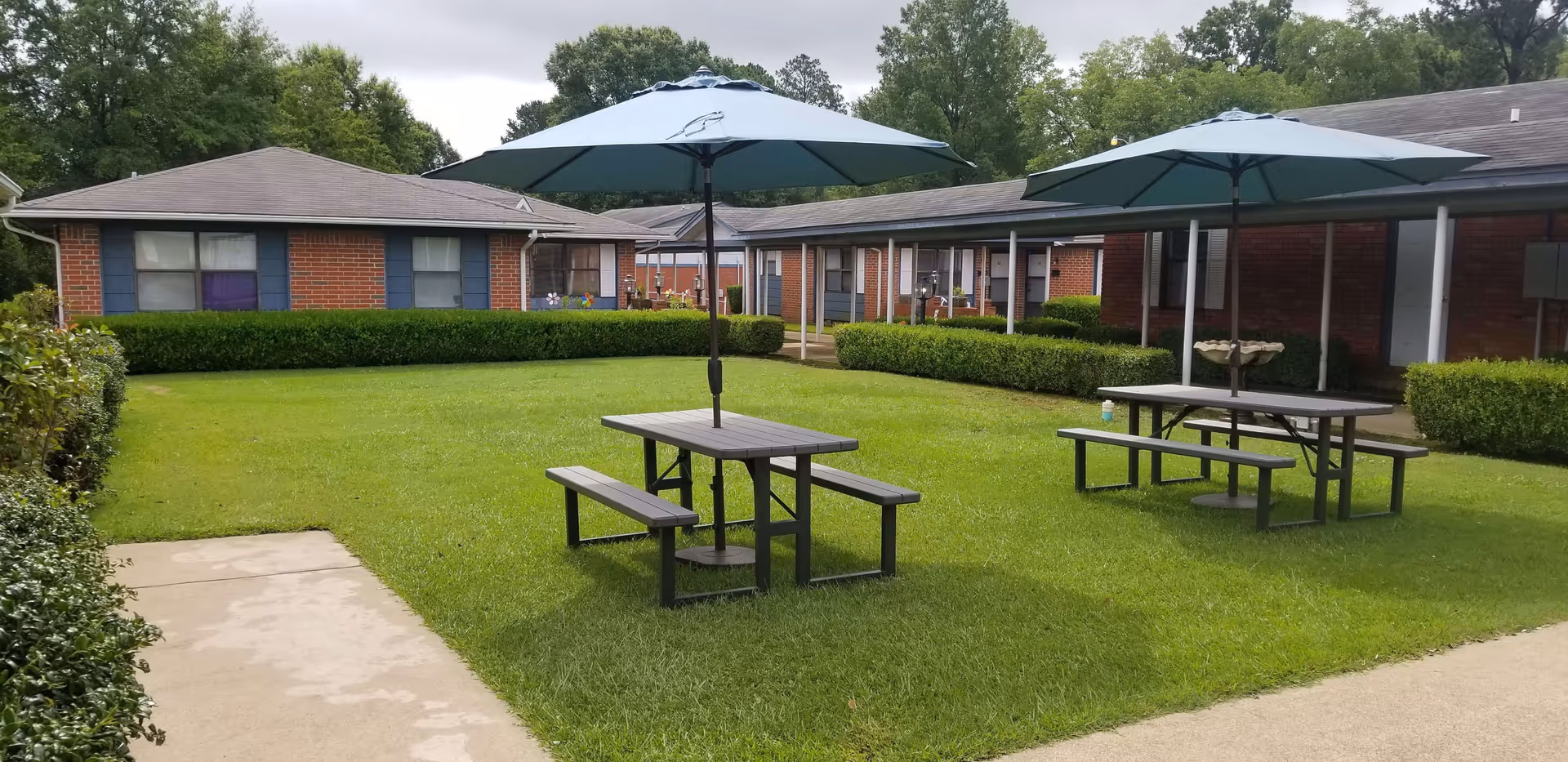 Outdoor courtyard area at Colonial Lodge Senior Living with green grass, two picnic tables each with a large blue umbrella, surrounded by single-story brick buildings with blue shutters and windows. There are neatly trimmed hedges and trees in the background under a cloudy sky.