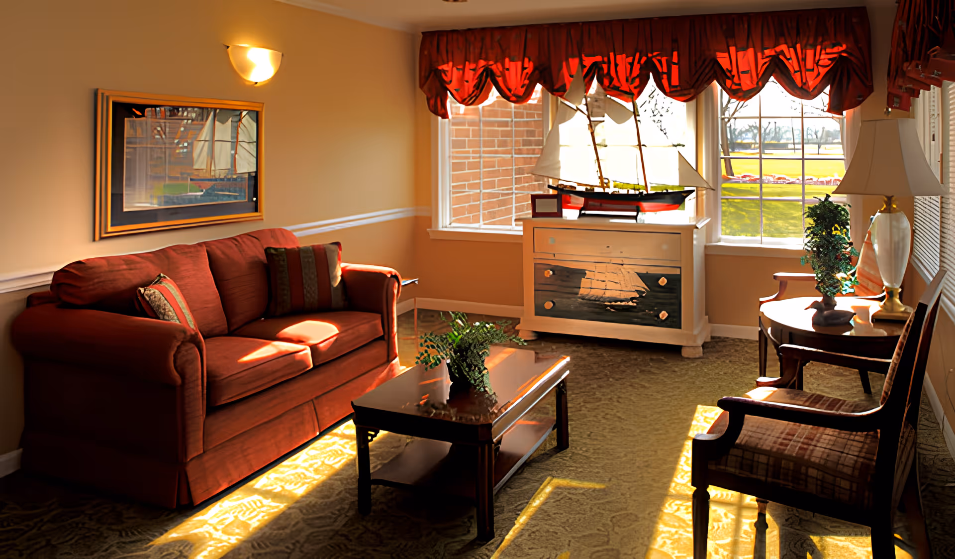 A cozy living room with a red sofa, two wooden chairs with cushions, a wooden coffee table with a plant, and a white cabinet with a model sailboat on top. The room has beige walls, a framed picture of a ship, red valance curtains on the windows, and sunlight streaming in, casting shadows on the carpeted floor.