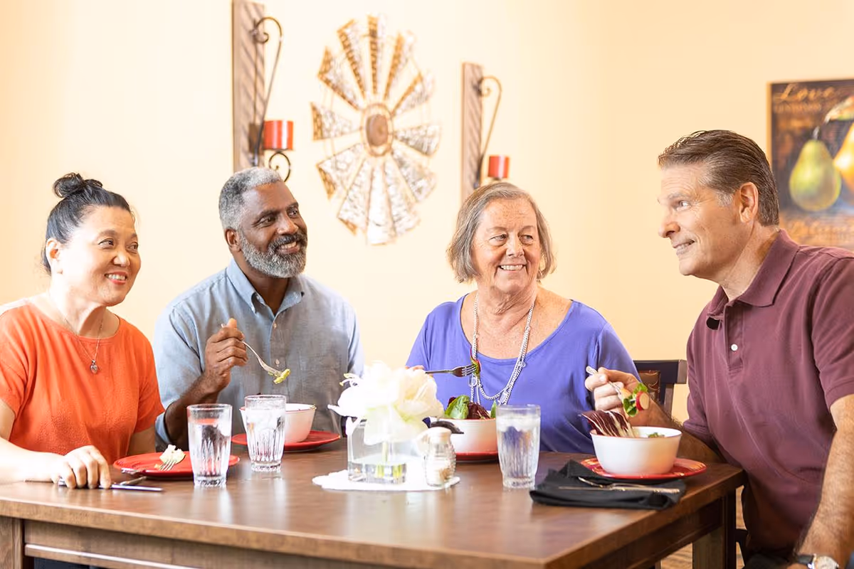 Four adults sitting around a dining table enjoying a meal together, smiling and engaging in conversation. The table is set with bowls of food, glasses of water, and a centerpiece with white flowers. The background features warm-colored walls with decorative wall art.
