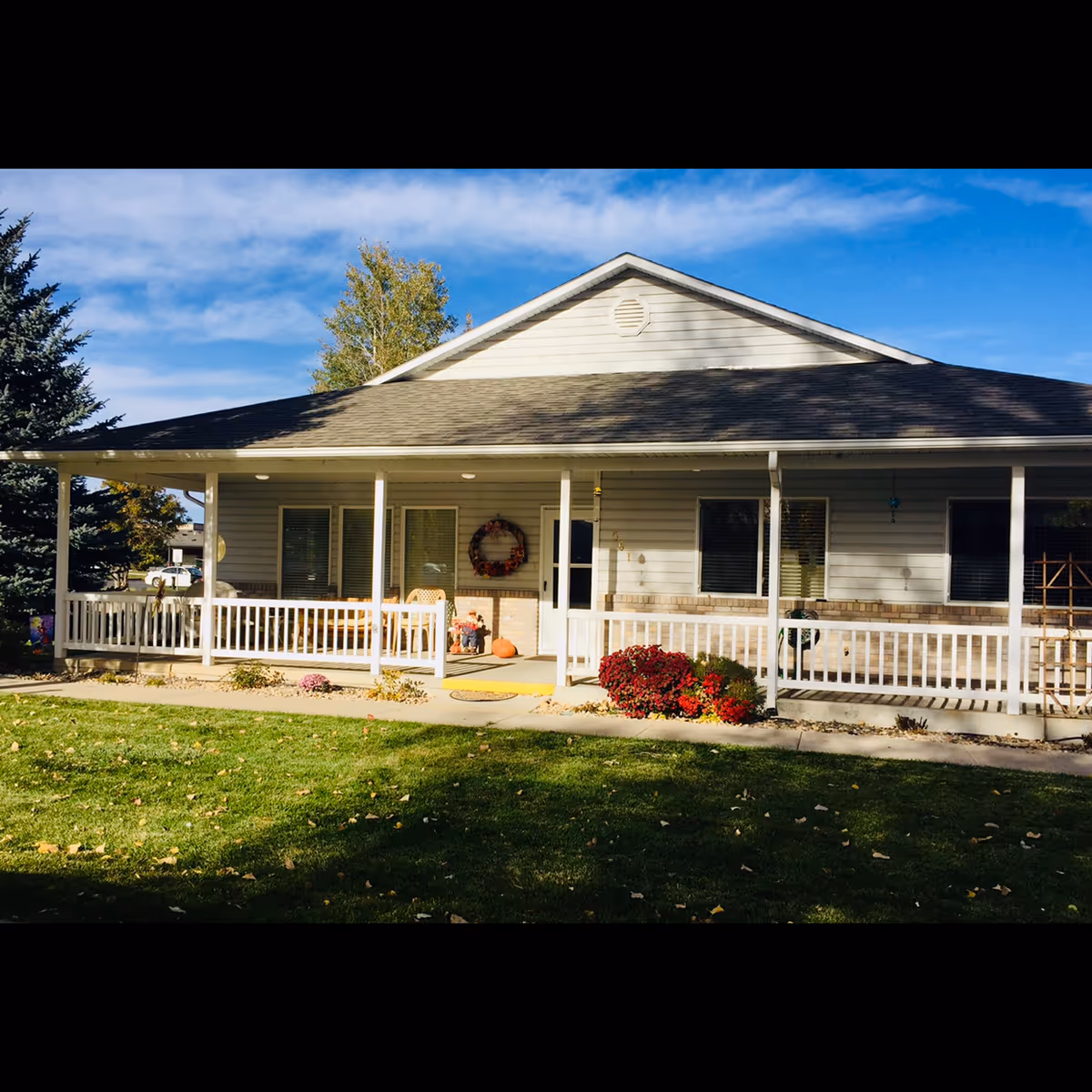 Single-story light-colored building with a covered front porch, white railing, seasonal decorations, and a green lawn.