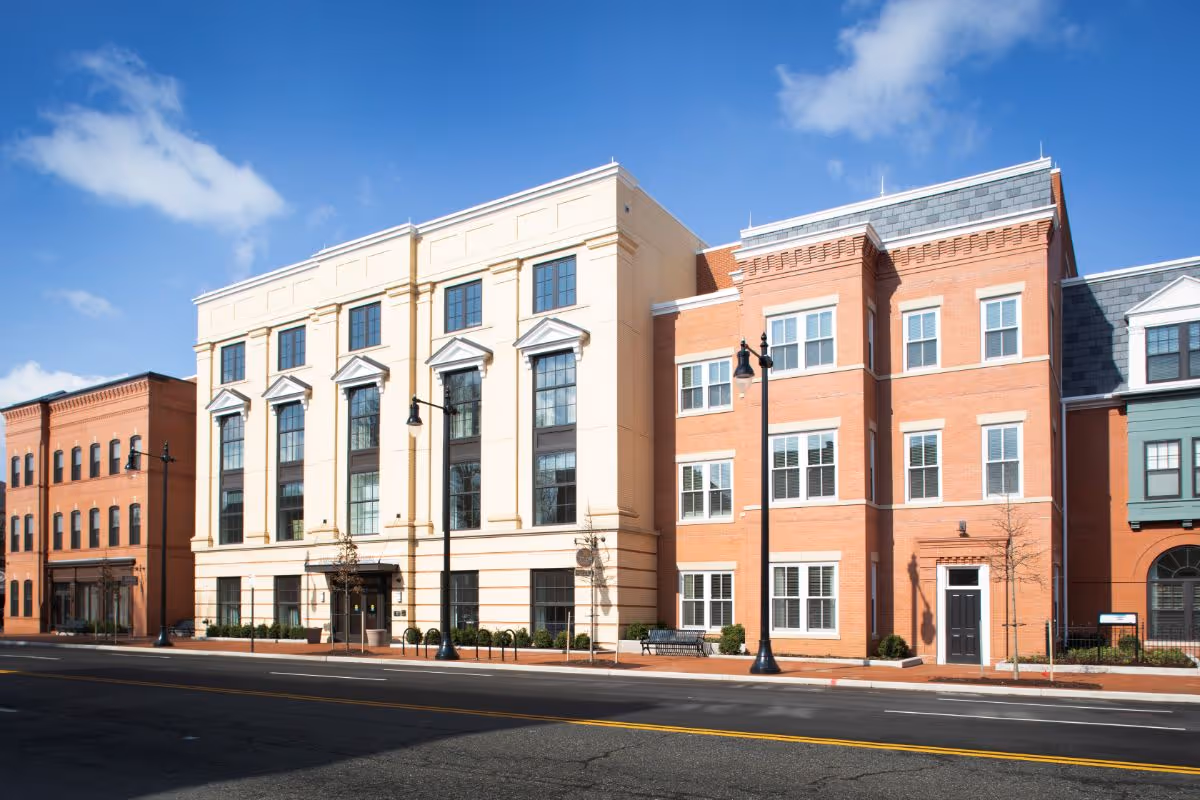 Exterior view of a multi-story senior living facility building with a combination of beige and red brick facades, large windows, street lamps, and a clear blue sky.