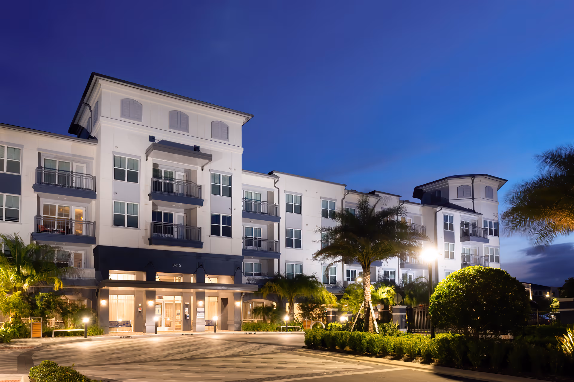 Illuminated multi-story senior living building with balconies, palm trees, and a landscaped entrance at dusk.