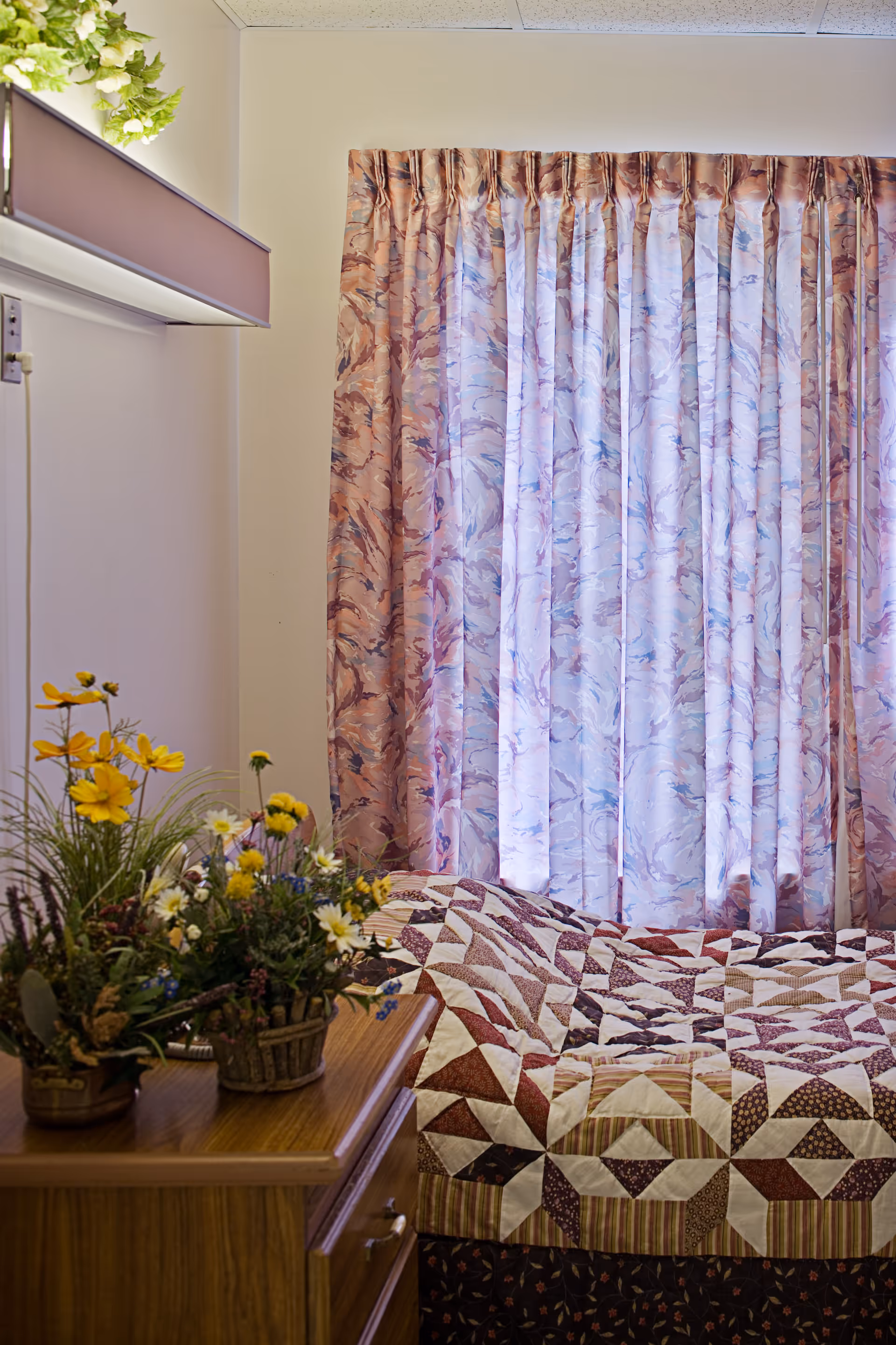 A cozy bedroom corner with a bed covered in a patchwork quilt featuring brown, beige, and white patterns. Next to the bed is a wooden nightstand with a floral arrangement in a basket. Behind the bed, there are pastel-colored curtains with a soft, abstract pattern covering the window. A small shelf with green plants is mounted on the wall above the nightstand.