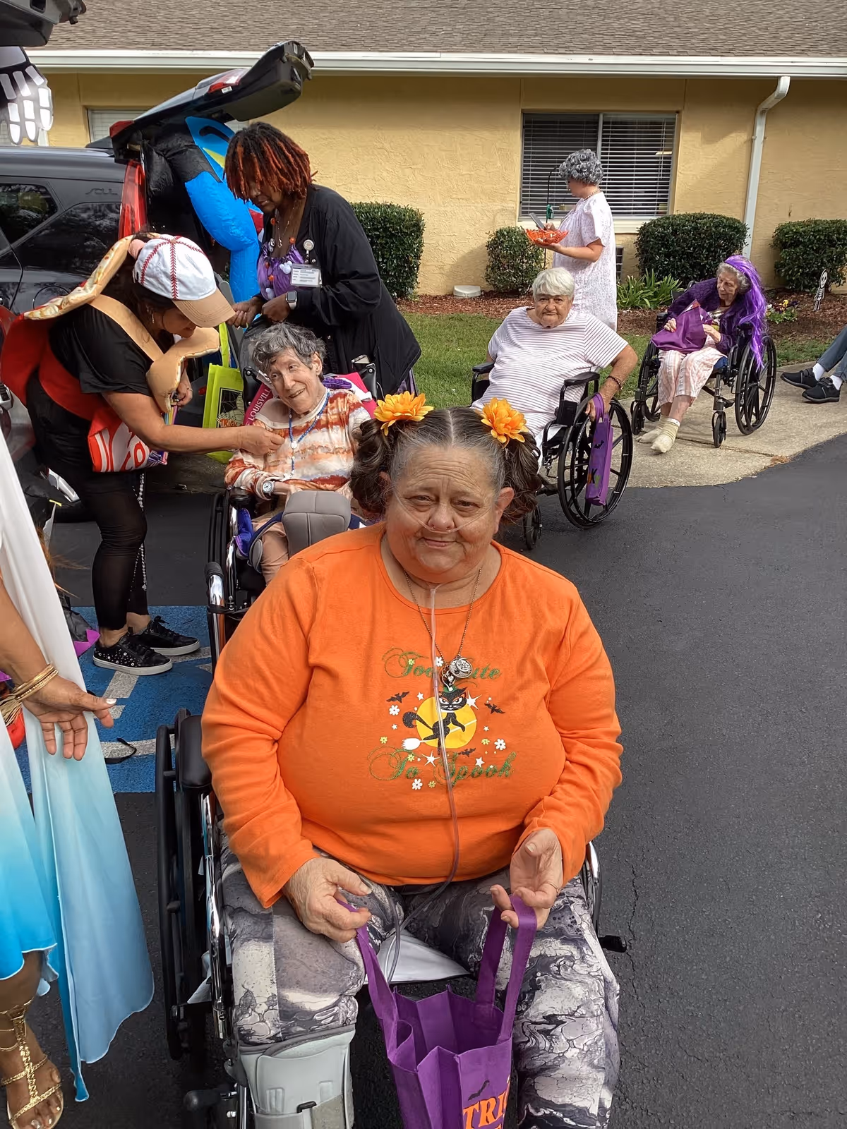 A group of elderly women, some in wheelchairs, are gathered outside near a building with yellow walls. One woman in the foreground wears an orange shirt with Halloween-themed decorations and has flowers in her hair. Other women and caregivers are interacting in the background, with some dressed in costumes. The scene appears to be a festive outdoor event at a senior living facility.
