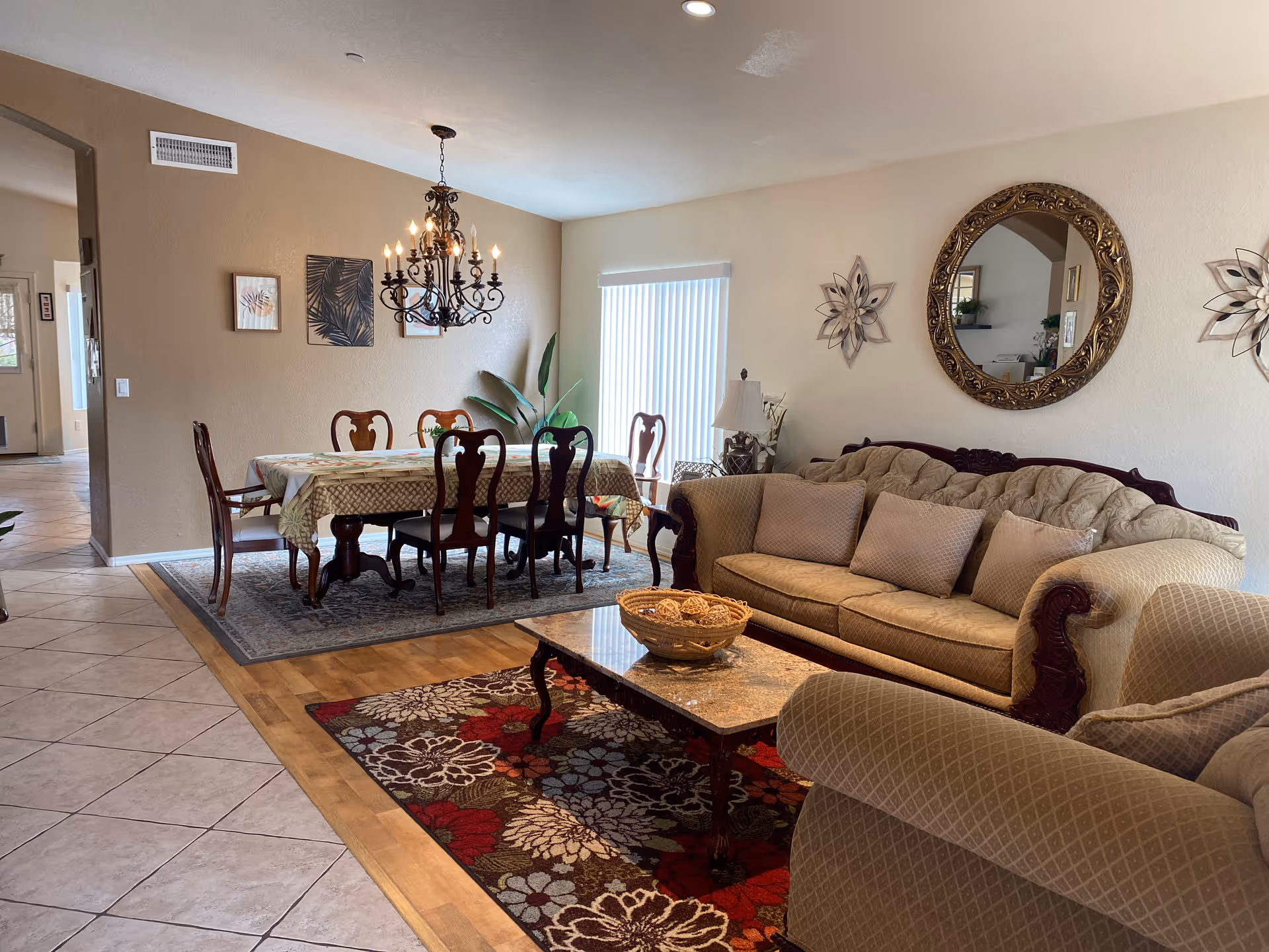 A cozy living and dining area with a beige sofa and armchair, a coffee table with a decorative bowl, and a dining table with six chairs covered with a patterned tablecloth. The room features a chandelier above the dining table, a large ornate round mirror on the wall, decorative wall art, and a sliding glass door with vertical blinds letting in natural light.