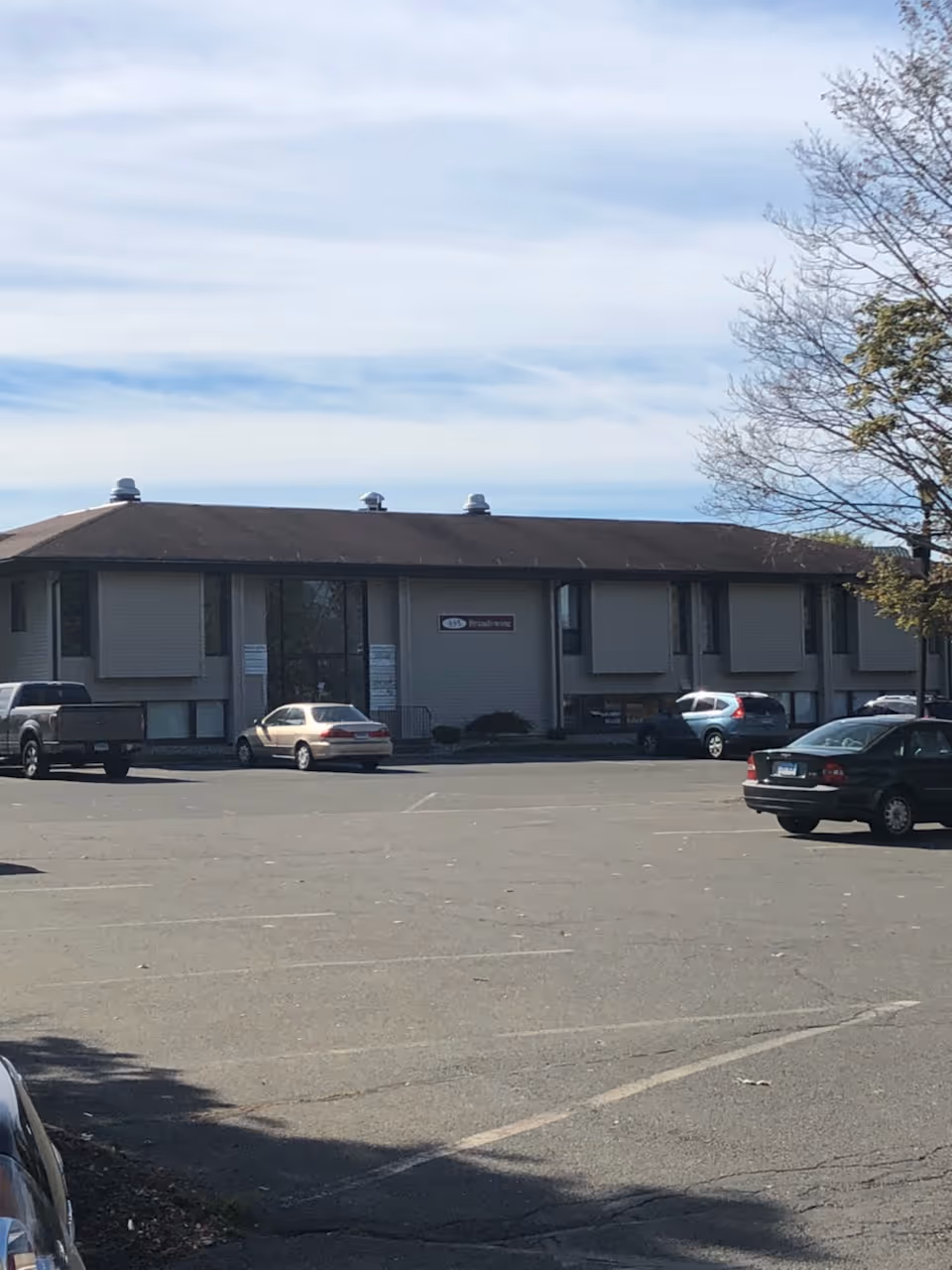 Two-story gray building with a wide parking lot and several parked cars.