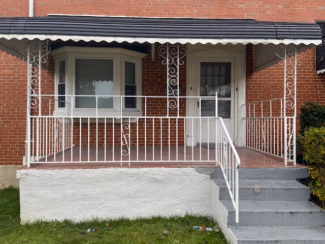 Brick building with a white wrought-iron porch railing, window, and front door.