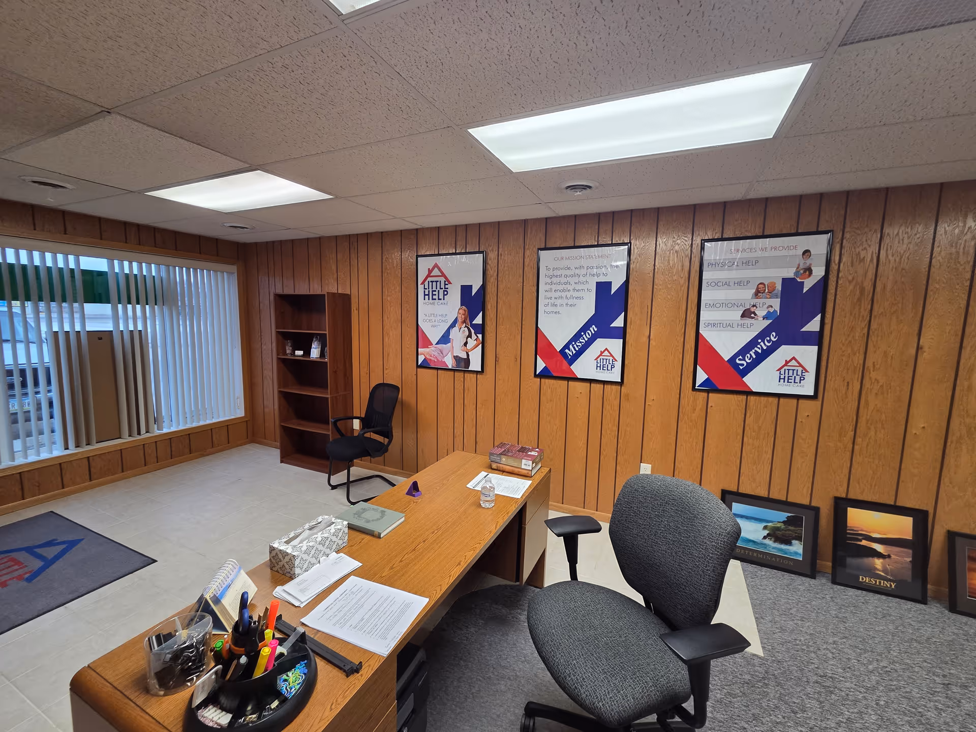 Wood-paneled office space with a desk, chairs, posters, a bookshelf, and a window with blinds.