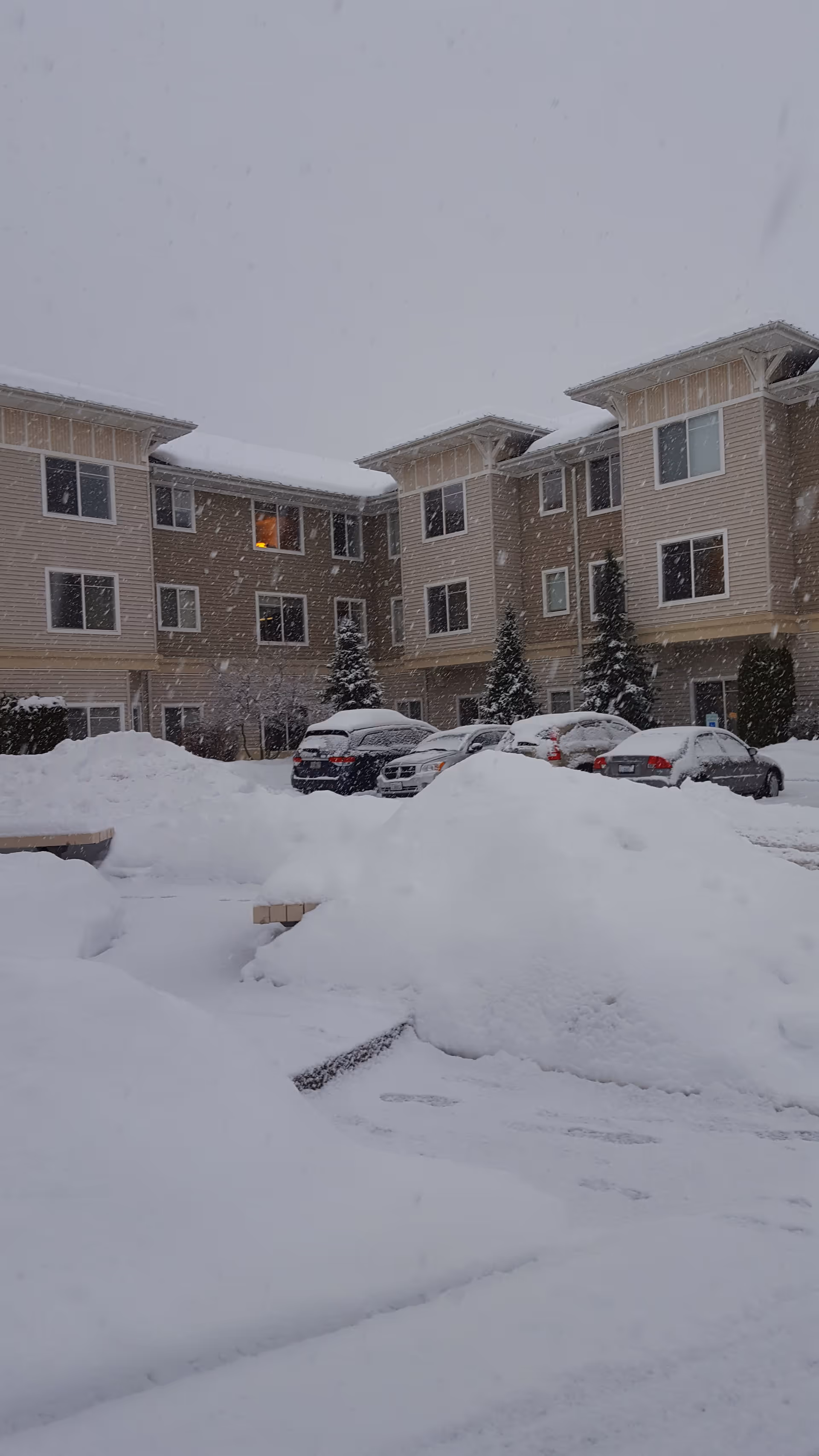 Snow blankets the front of a multi-story senior living building with parked cars.