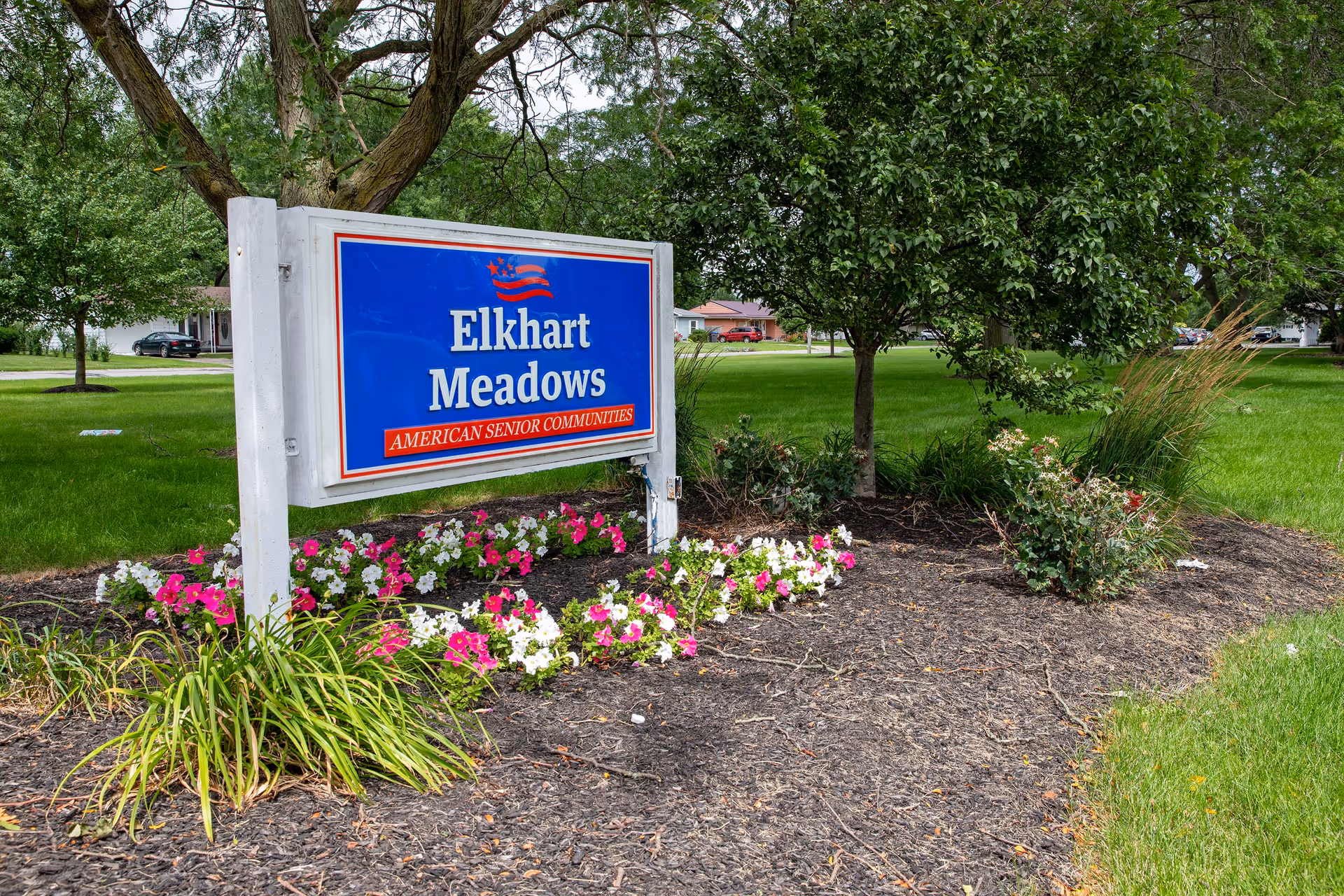 A blue and white sign for Elkhart Meadows American Senior Communities is displayed outdoors in a landscaped area with pink and white flowers, green grass, and trees in the background.