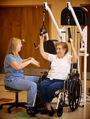 A healthcare worker in blue scrubs assists an elderly woman in a wheelchair as she uses an exercise machine with pulleys in a room with wooden doors.