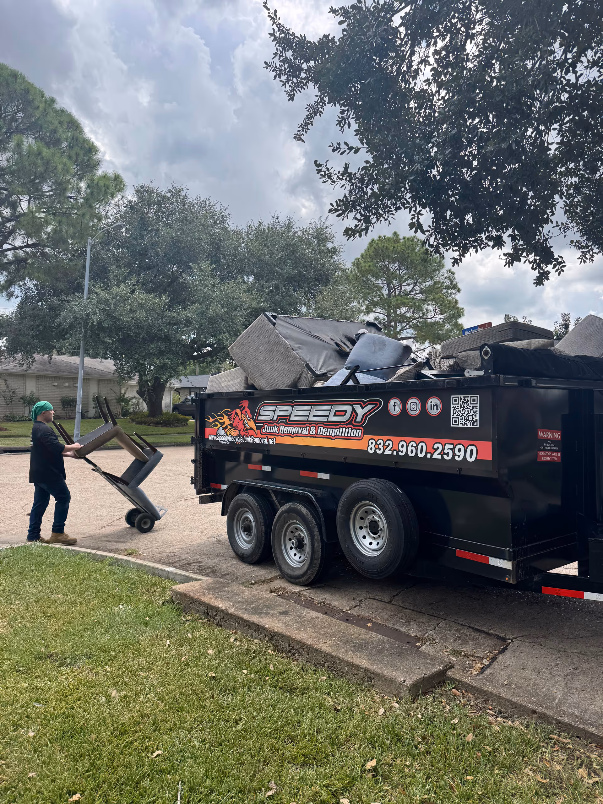 A worker pushing a hand truck loaded with chairs toward a large junk removal trailer outdoors.