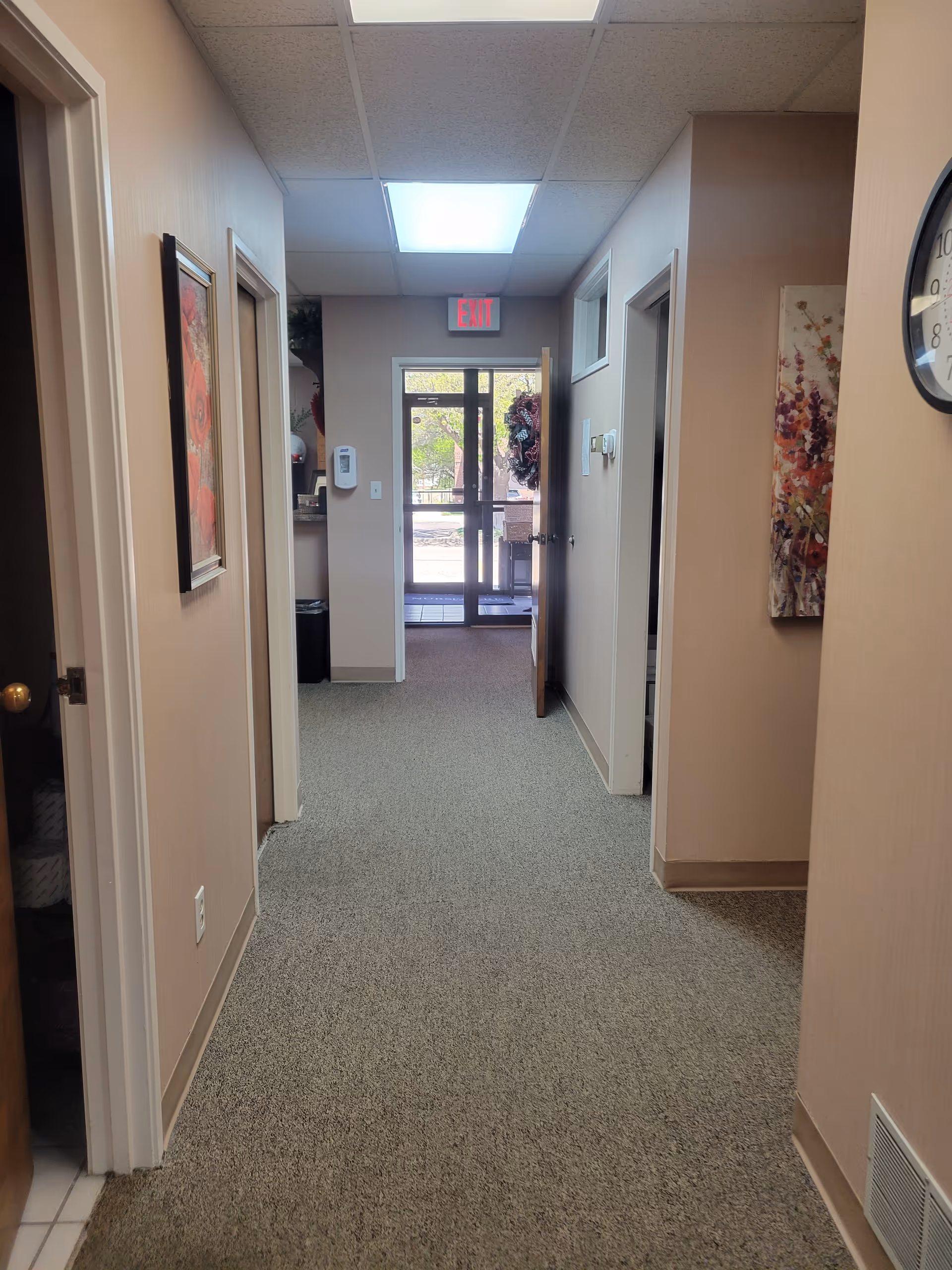 A hallway in a senior living facility with doors, artwork, and an exterior exit at end.