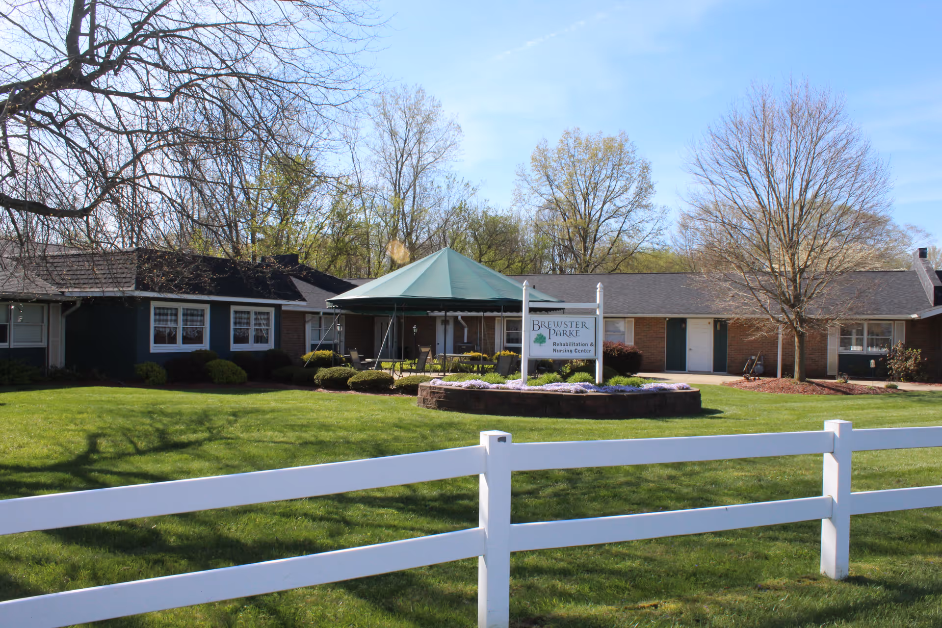 Exterior view of Brewster Parke, Inc, a rehabilitation and nursing center, showing a single-story building with a green canopy over an outdoor seating area, surrounded by green grass, trees, and a white fence in the foreground.