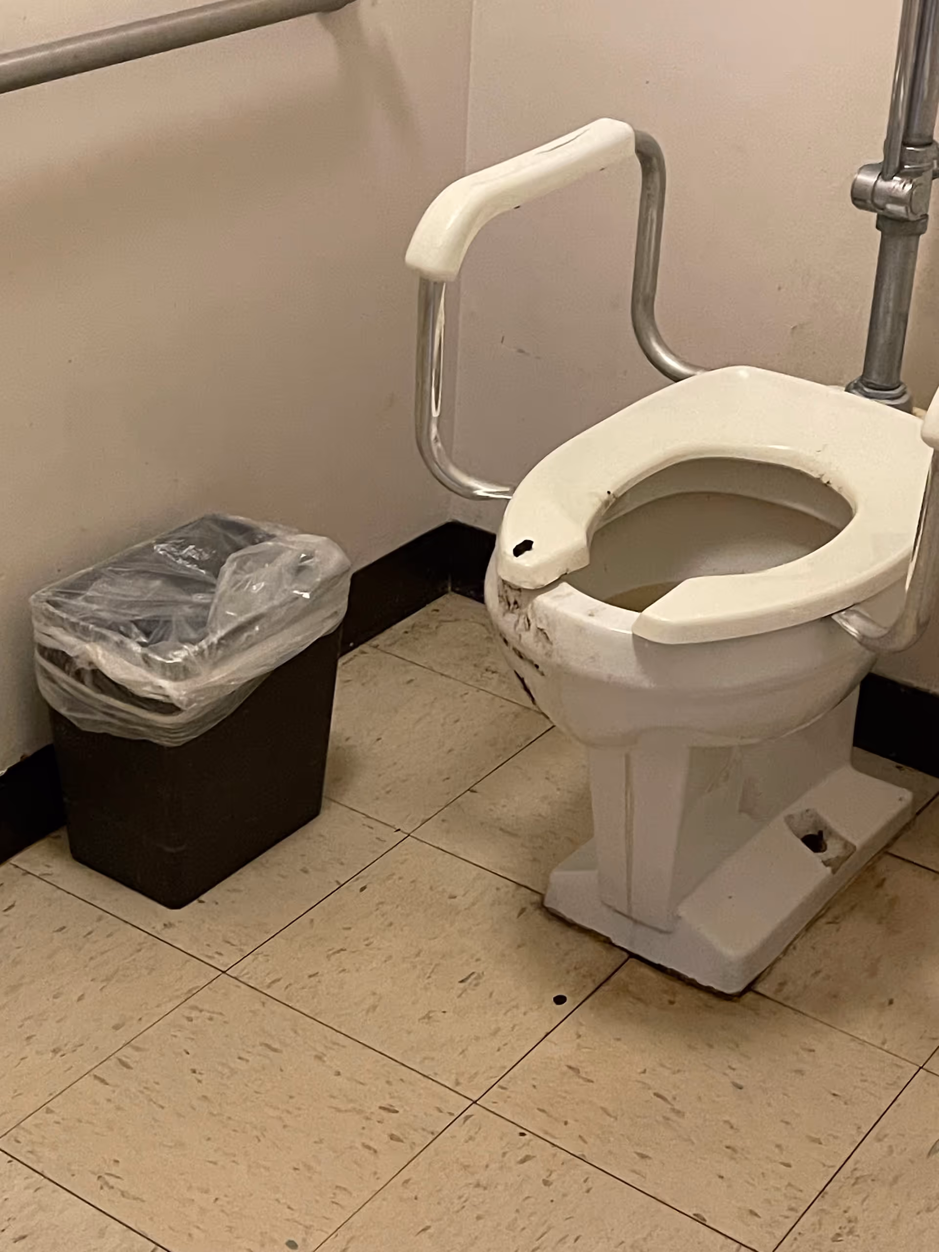 Accessible hospital-style bathroom with a raised toilet seat, safety rails, and a trash can.