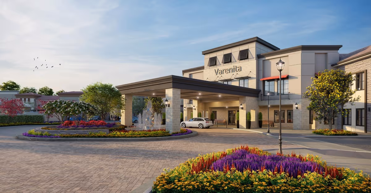 Front entrance of the Varenita senior living building with a covered porte-cochere, fountain, landscaped flowerbeds and a parked car.