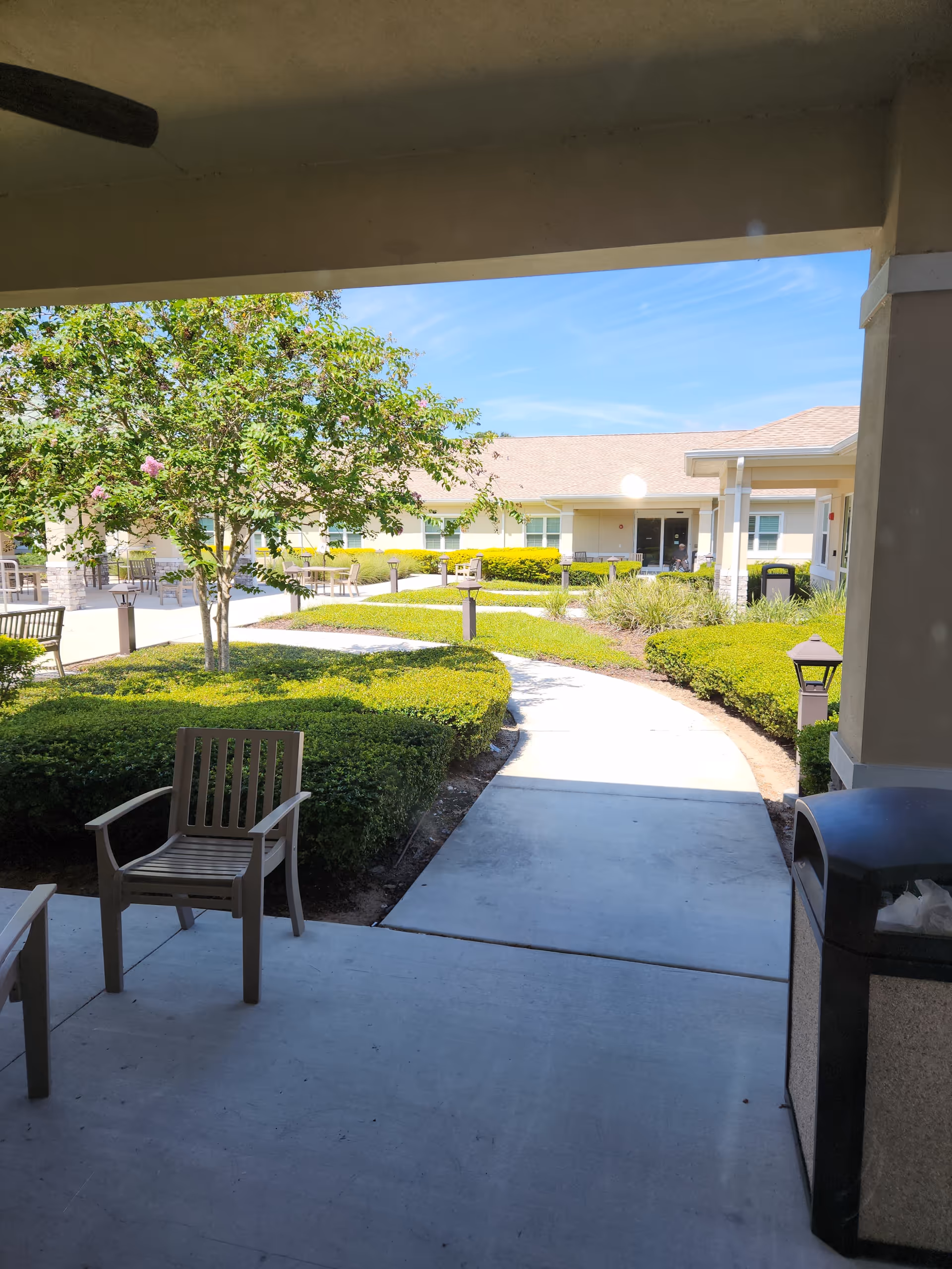 Outdoor courtyard of a senior living center with paths, greenery, benches, and shaded seating.