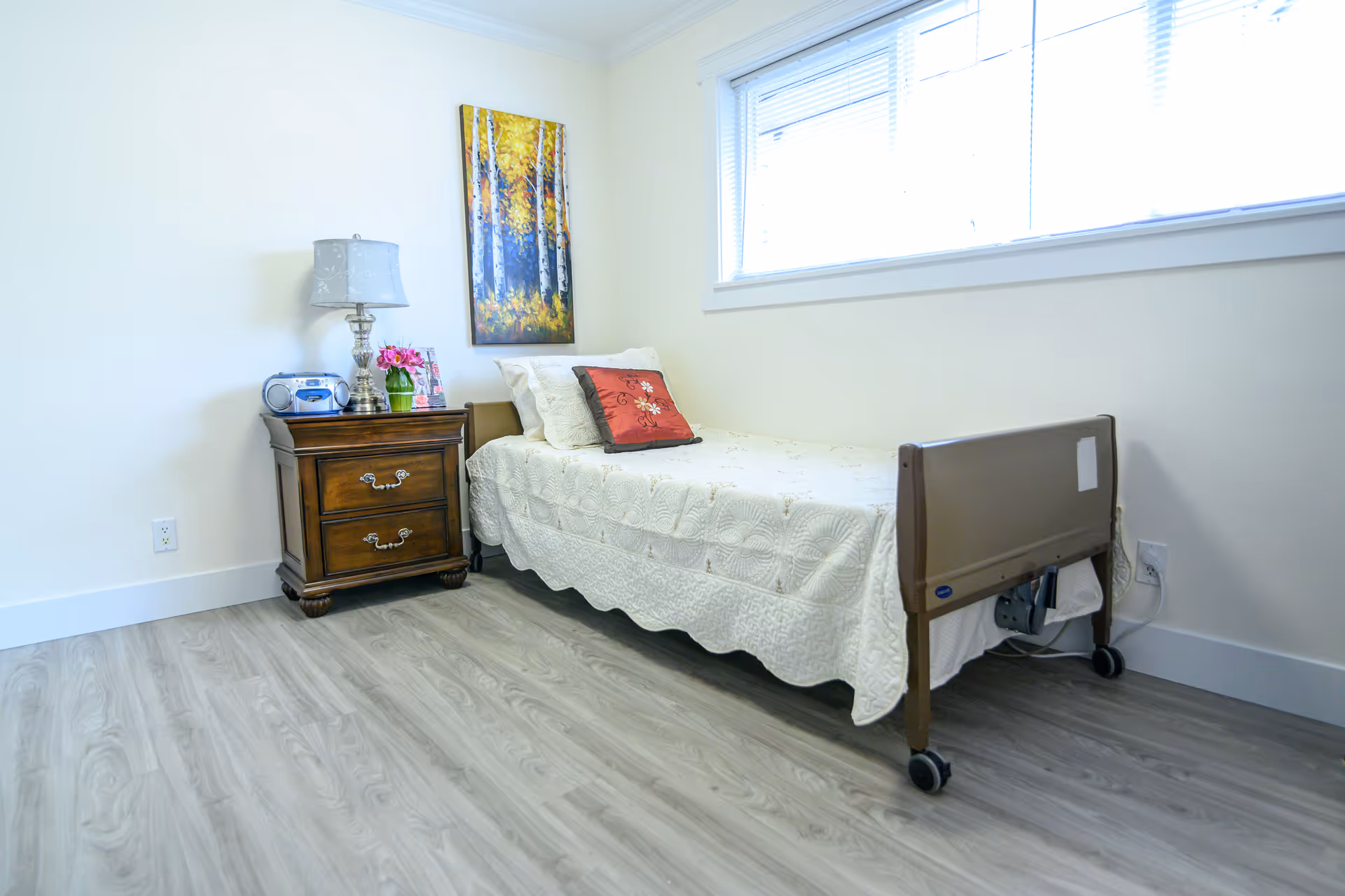A single bed with white quilt, nightstand, lamp, and window in a bright bedroom.