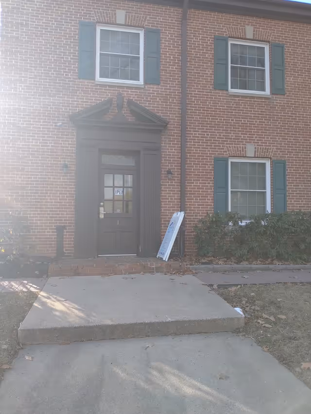 Brick building entrance with dark door, green shutters, and a sidewalk signboard.