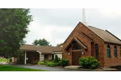 Brick building with arched entrance, shrubs, and a carport on a sunny day.