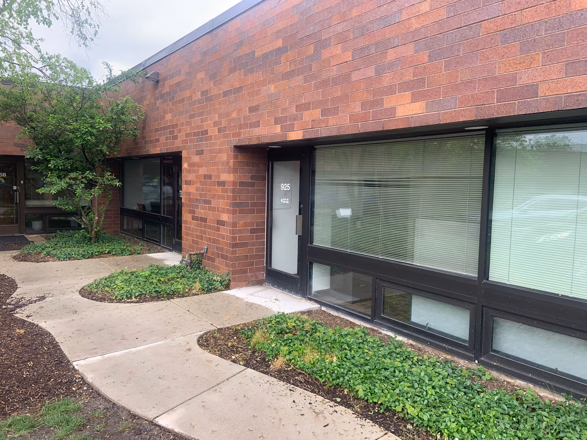 Brick facility exterior with entrance door, large windows, and surrounding landscaping.